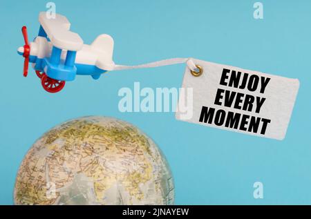 Travel and business concept. On a blue background, a globe and an airplane with a sign - Enjoy every moment. Globe out of focus. Stock Photo