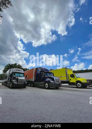 Three large, colorful trucks parked on asphalt at a highway rest stop under a bright blue sky with dramatic clouds.l Stock Photo