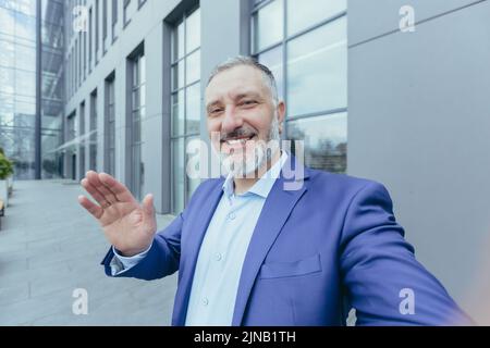 Senior successful and experienced gray haired man looking into phone camera and smiling talking on video call, businessman taking selfie while standing outside office building Stock Photo