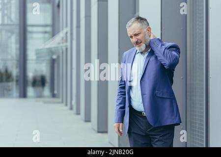 Injured and overtired senior gray-haired businessman, man outside office has severe neck pain, director holding hand massaging neck Stock Photo