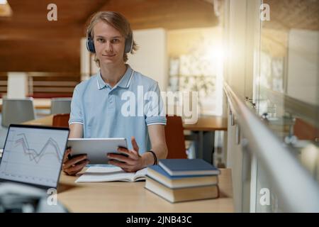 Smiling Male student sits in headphones in library and studies using a digital tablet Stock Photo