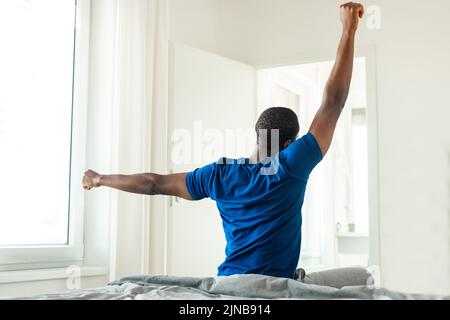 Black Male Stretching Arms Sitting Back To Camera In Bedroom Stock Photo