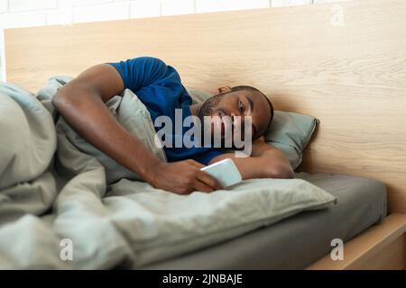 African American Guy Using Phone Texting Lying In Bedroom Indoor Stock Photo