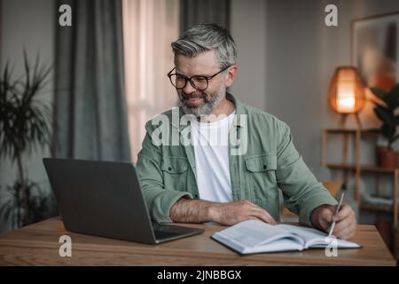 Glad senior european male with beard in glasses watches video on computer, makes notes at table in room Stock Photo