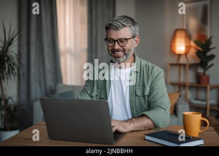 Cheerful elderly european man with beard in glasses watches video lesson, chatting on computer Stock Photo