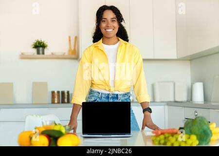 Cooking recipes online. Happy black lady showing laptop with blank screen in modern kitchen, mockup, copy space Stock Photo