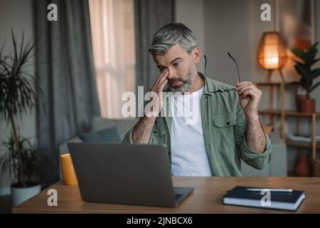 Sad tired retired caucasian man with beard takes off his glasses, rubs eyes looks at computer in room interior Stock Photo