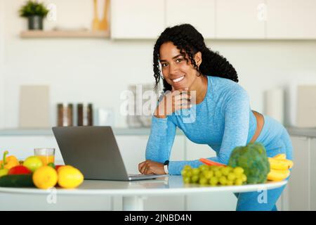 Searching for new healthy recipes. Happy fit lady cooking fresh salad at home in modern kitchen, using laptop Stock Photo
