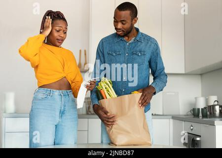 Shocked black spouses with paper bag with food checking grocery bill, woman touching her head, standing in kitchen Stock Photo