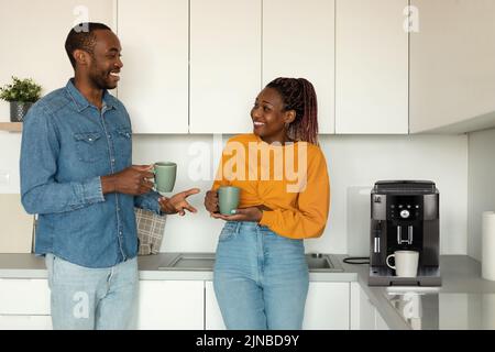 Young african american spouses drinking morning coffee and talking while standing near coffee machine in kitchen Stock Photo