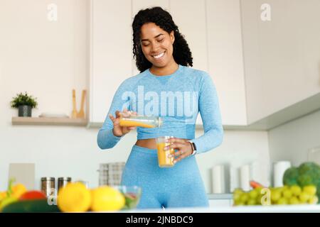 Happy black fit woman having fresh juice for breakfast in the kitchen, pouring beverage from bottle to glass Stock Photo