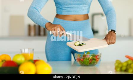 Detox recipes. Fit black lady cooking healthy vegetable salad, cutting cucumber on wooden board, cropped Stock Photo