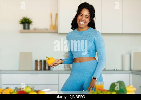 Happy fit black lady holding bell pepper and smiling at camera, cooking dieting dinner in kitchen interior Stock Photo
