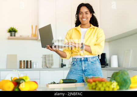 Happy black woman holding and using laptop, searching for new recipes, cooking healthy meal in modern kitchen Stock Photo