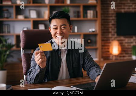 Satisfied middle aged chinese man at table with laptop use credit card in home office interior Stock Photo