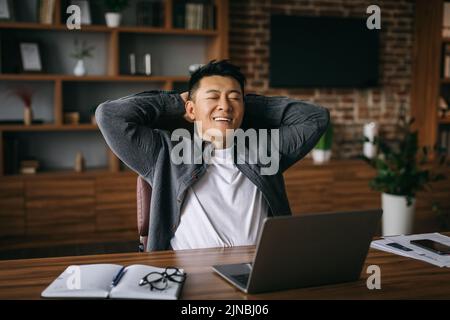 Smiling adult chinese male enjoys rest in chair at table with computer in office interior Stock Photo