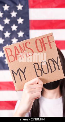 Young woman holding sign Bans Of My Body covering face stop abortion American flag on background. Anti domestic violence, racism racial discrimination Stock Photo