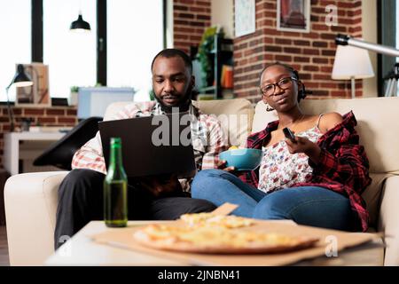 Boyfriend working on laptop and girlfriend switching tv channel, eating takeout delivery meal and watching movie on television. Enjoying fast food and beer, using computer at home. Stock Photo
