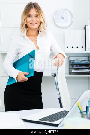 Friendly girl with blue folder of documents stands in bright office Stock Photo