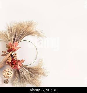 Floral wreath from dry pampas grass and Autumn leaves. Hands tie decorations to metal frame. Stock Photo