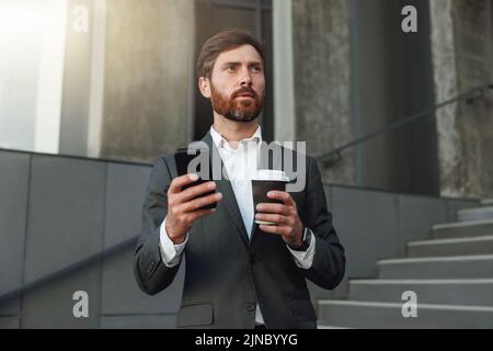 Focused office worker in suit standing on stairs with coffee during break and using phone Stock Photo