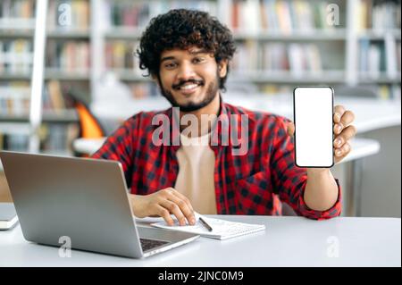 Defocused Indian or Arabian male student, sitting at table in library, showing his smart phone with blank white screen for advertising and presentation, smile friendly. Mock-up and copy-space concept Stock Photo