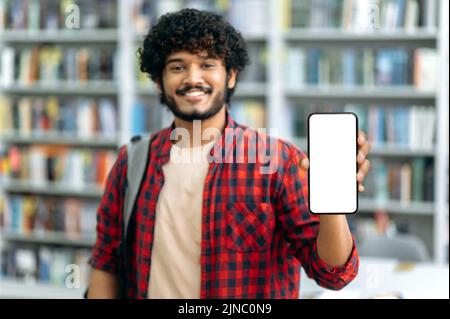 Gadgets, mock-up concept. Defocused positive attractive indian or arabian guy, freelancer or student, showing his smartphone with blank white mock-up screen, looks at camera, smiles happily Stock Photo