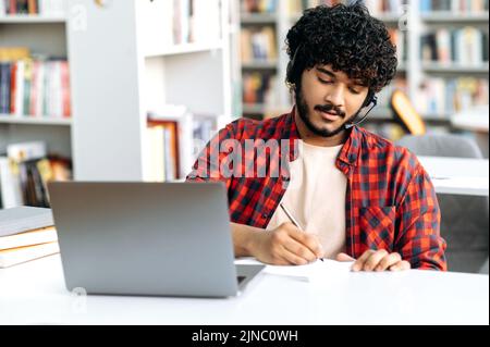 Focused clever arabian or indian male student, with headset, sits at table with laptop in university library, preparing for exam, studying information, listens lecture, concentrated takes notes Stock Photo
