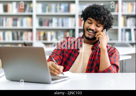 Phone talk conversation during work or study. Positive busy indian or arabian curly haired happy guy, having negotiation by smart phone, talks with friend or colleague, sits at desk, smiling friendly Stock Photo