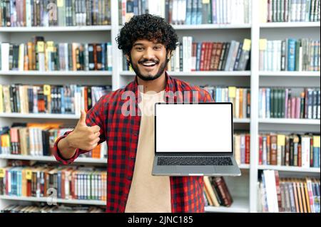 Excited millennial arabian or indian male student, shows a laptop with blank white mock-up screen, stands in a library against the background of bookshelves, looks at camera, smiles, thumb up gesture Stock Photo
