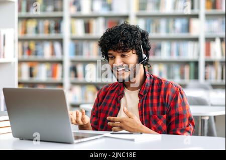 Friendly stylish Indian or Arabian guy, male student or freelancer with headset, sitting at a desk in a library, using laptop for online conversation, discuss information with interlocutor, smiling Stock Photo