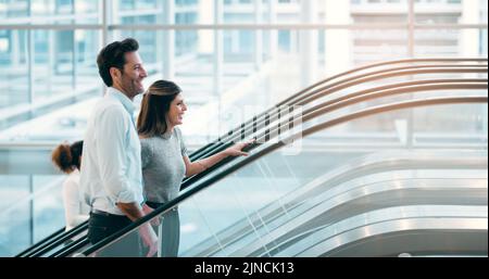 Theyre excited about working together. two young businesspeople going up an escalator in a modern workplace. Stock Photo