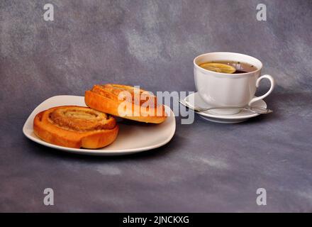 Two sweet rolls with sugar and cinnamon rolled into a spiral on a gray background, next to a cup of tea with lemon. Close-up. Stock Photo