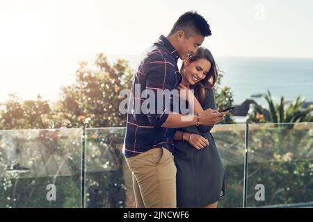 We love sharing our best moments online. an affectionate young couple using a cellphone together while relaxing outdoors. Stock Photo