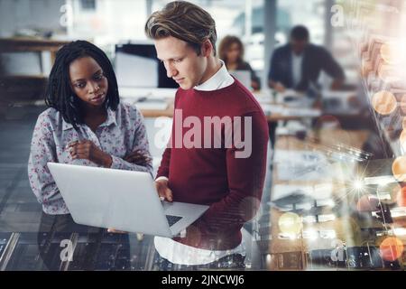 Creative juices flowing all around the office. two young businesspeople using a laptop together with their colleagues working in the background at Stock Photo