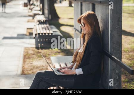 Side view of a ginger business woman freelancer sitting on bench working with laptop in city park. A beautiful happy woman using laptop outdoors Stock Photo