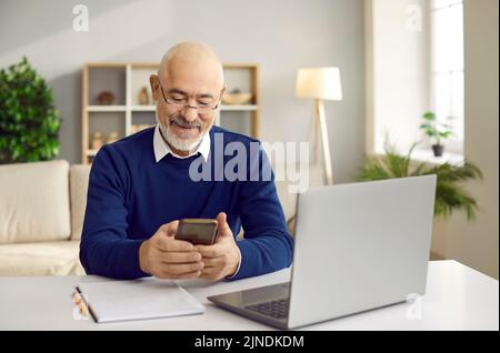 Happy senior man using his mobile phone while sitting at desk with laptop computer Stock Photo