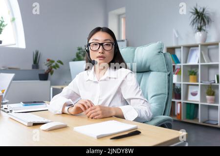 Asian woman in headphones with a microphone, sitting in the office at the desk conducting an online webinar, explaining, consulting, telling, gesturing with her hands. He looks at the camera, smiles. Stock Photo
