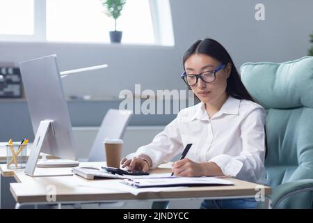 Workaholic. A beautiful young Asian business woman in glasses is very early at work, sitting at the table drinking coffee to go, a paper cup of coffee is standing on the desktop near the computer. Stock Photo