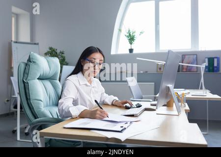 Work with documents. Portrait of a young beautiful business woman Asian accountant works with documents and reports. Sitting at the desk in the office, writing. Stock Photo