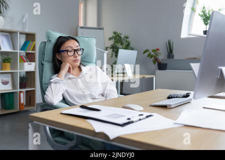 Pensive and sad young Asian businesswoman sitting in the office at the desk in a big chair, thinking about solving a problem Stock Photo