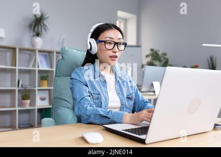 Online training. Young beautiful Asian girl student in glasses studying at a laptop in headphones. Learns remotely, sits at a desk in a modern office. Stock Photo