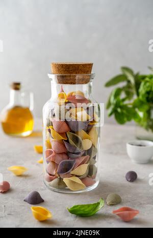Multi-coloured Italian pasta conchiglie or seashells in glass bottle, olive oil, olive branch. Concrete background  Stock Photo
