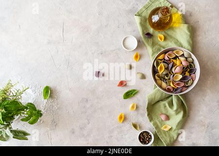 Multi-coloured Italian pasta conchiglie or seashells in bowl, olive oil, spices, herbs, green napkin. Flatly with copy space.  Stock Photo
