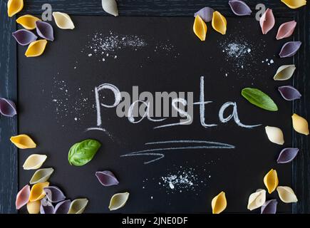 Multi-coloured Italian pasta conchiglie or seashells with basil leaves on chalk board. Word Pasta wrote with chalk Stock Photo