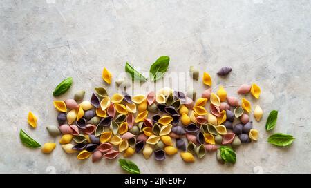 Multi-coloured Italian pasta conchiglie or seashells with basil leaves on concrete background. Flatly with copy space.  Stock Photo