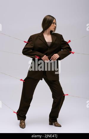 full length of teenage model in stylish brown suit pinned with clothespins on rope posing on grey Stock Photo