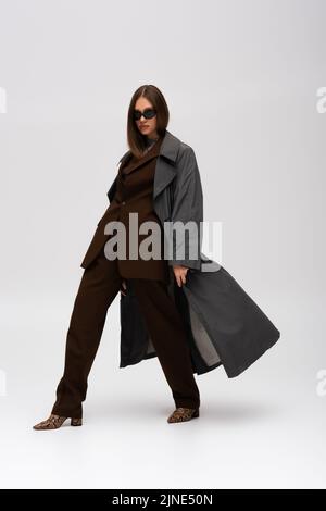 full length of stylish teenage model in sunglasses and trendy trench coat walking on grey Stock Photo