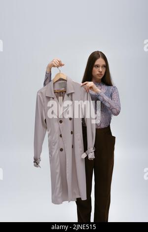 teenage model holding recyclable trench coat on hanger isolated on grey Stock Photo