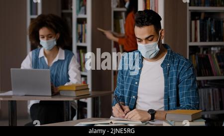 Students study in library focused intelligent young indian guy in protective mask sits at desk doing homework preparing for exam writes notes in Stock Photo